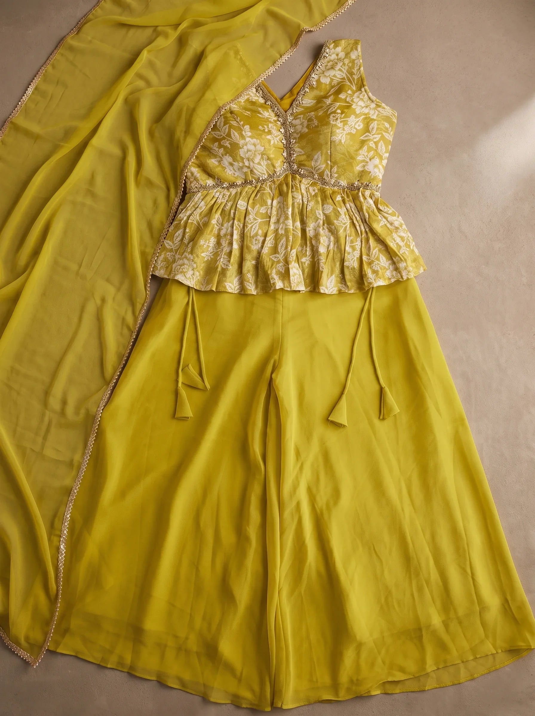 Flat lay of mustard yellow georgette floral peplum top, flared sharara bottoms and matching bordered dupatta