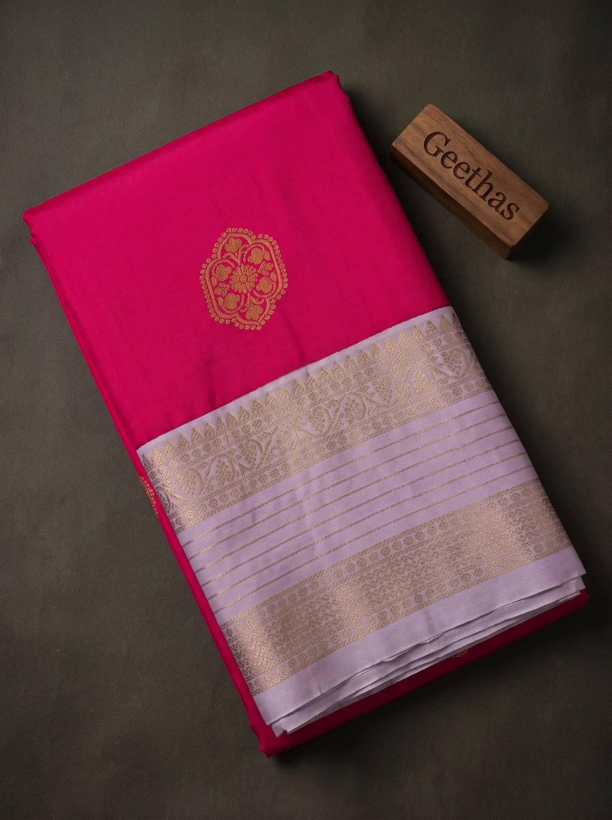 Folded magenta soft silk saree showing a gold-toned floral motif and lavender woven zari border