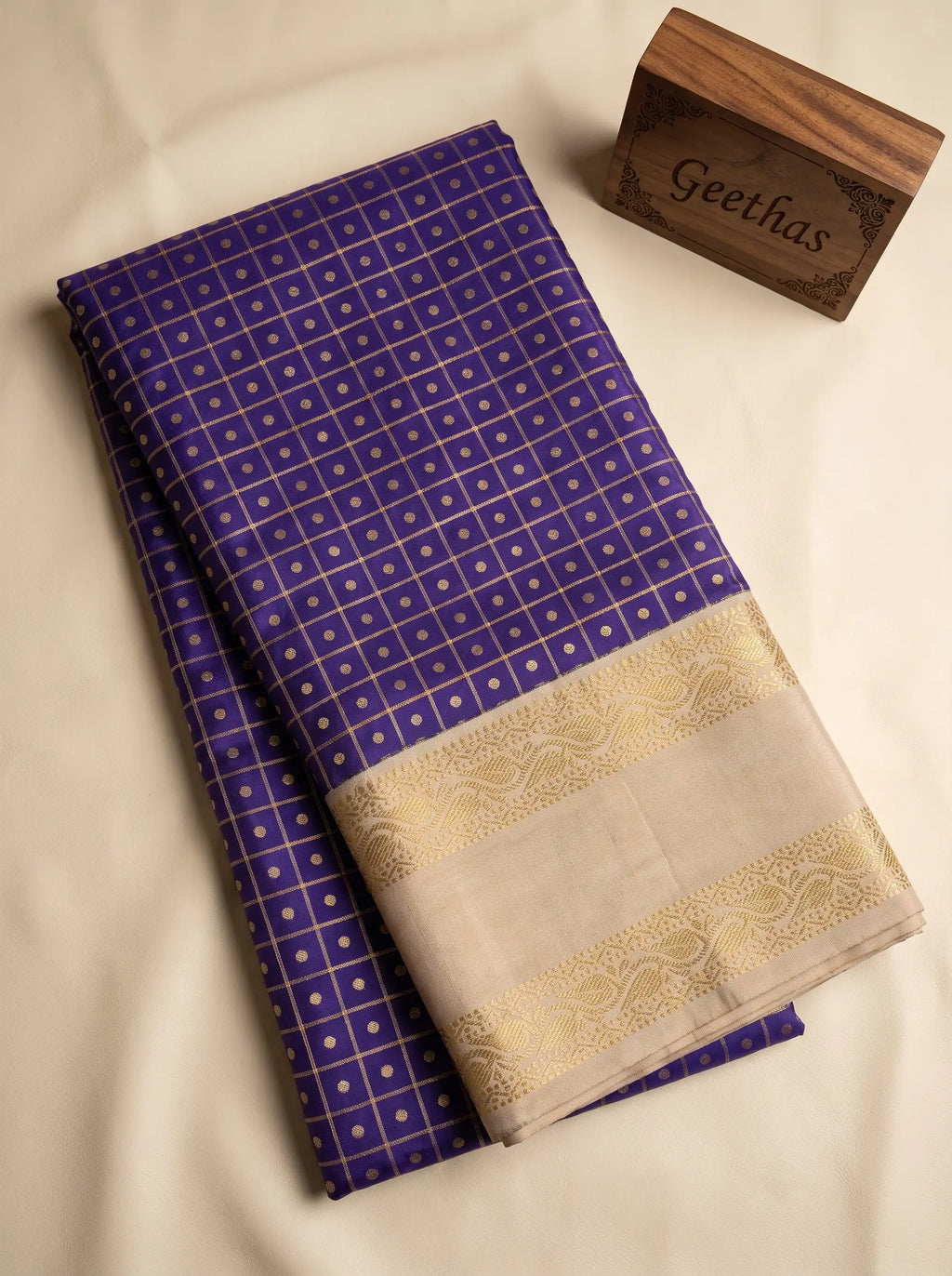 Folded violet soft silk saree showing gold geometric grid motifs and a cream decorative woven border.
